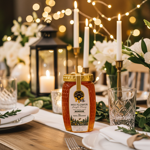 A jar of Sweetener's Mix Flower Honey displayed on a rustic wooden table, surrounded by a beautifully set dining table with white flowers, lit candles, and warm bokeh string lights in the background.