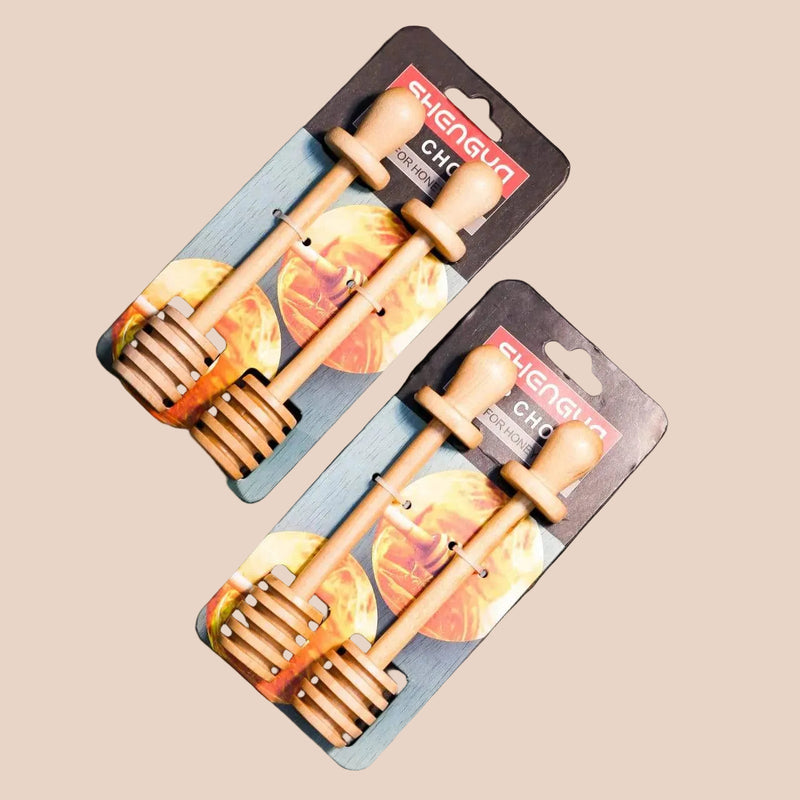 Two packaged wooden honey dipper spoons on a dark wood surface. The spoons are light natural wood with a slotted head, attached to blue backing cards with orange and yellow graphics and the brand name SUGARUN visible.